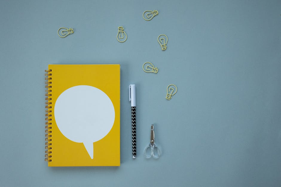Creative workspace setup with a yellow spiral notebook, pen, scissors, and light bulb paper clips on a blue background.